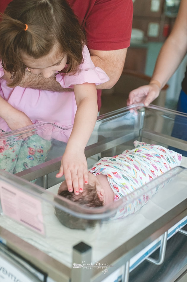 Dad holding big sister up so that she can reach in and touch her newborn baby sister sleeping in the bassinet Methodist Le Bonheur Germantown Hospital