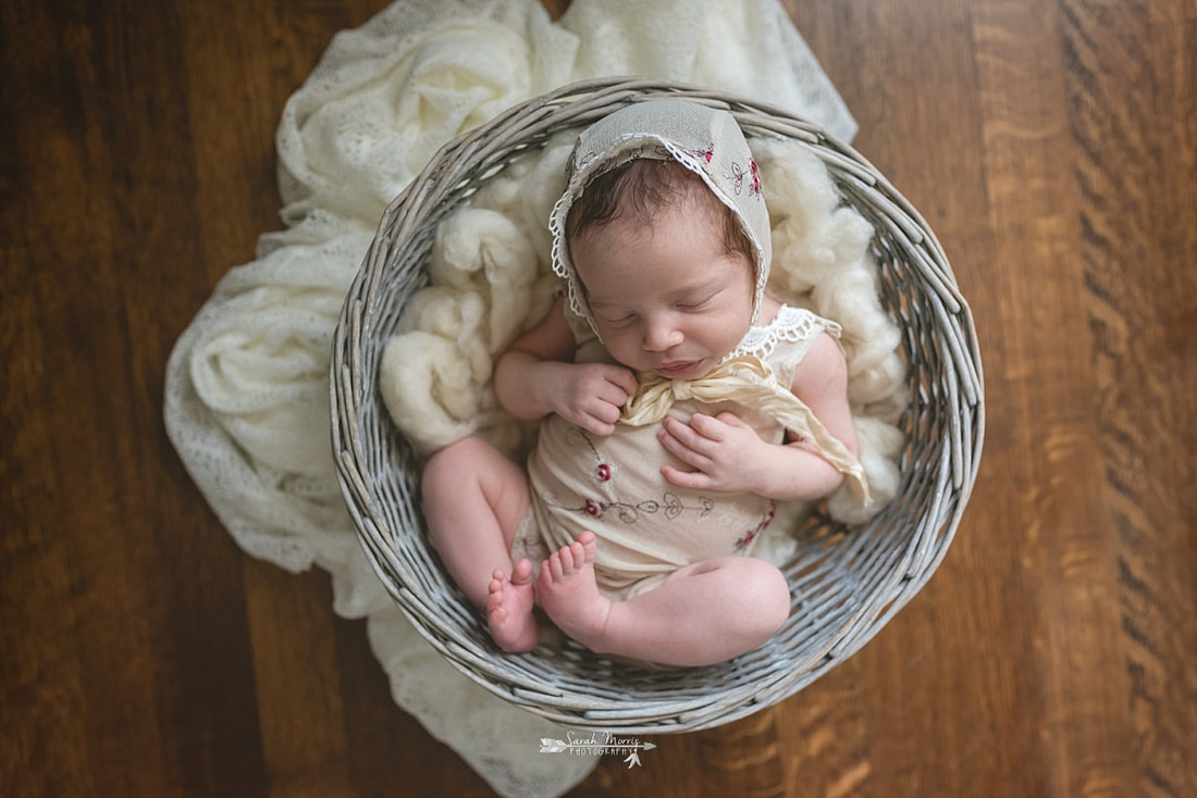 Newborn baby girl, wearing a floral romper and matching bonnet, sleeping in a basket on a wood backdrop