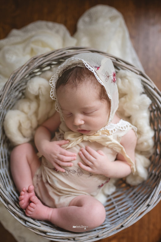 Newborn baby girl, wearing a floral romper and matching bonnet, sleeping in a basket on a wood backdrop