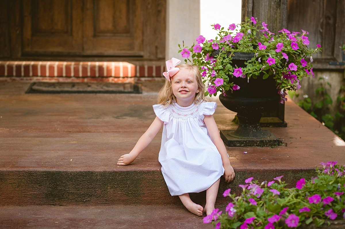 little girl wearing a smocked dress on the front porch by purple petunias during in home lifestyle family session in collierville, tn