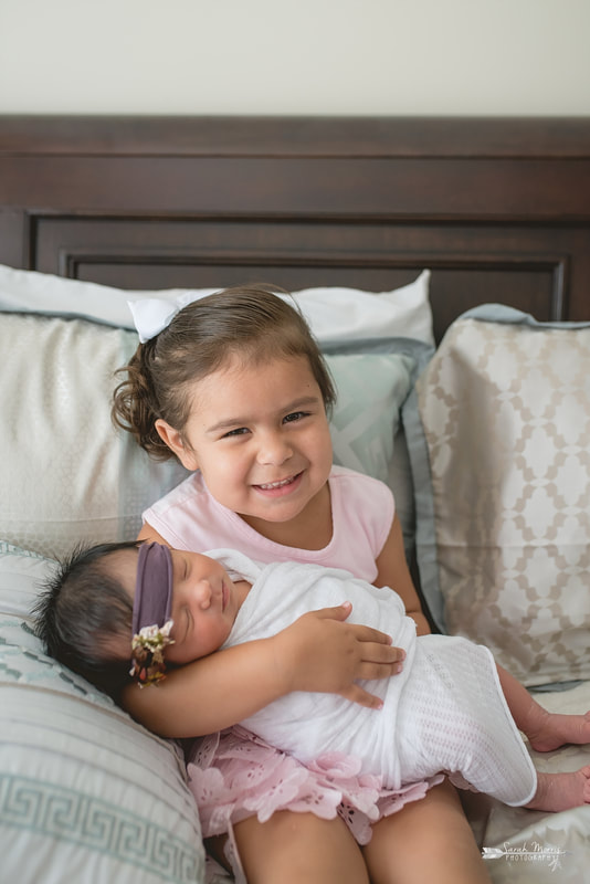Big sister holding baby sister on bed with newborn baby sister for the lifestyle portion of her newborn photo session