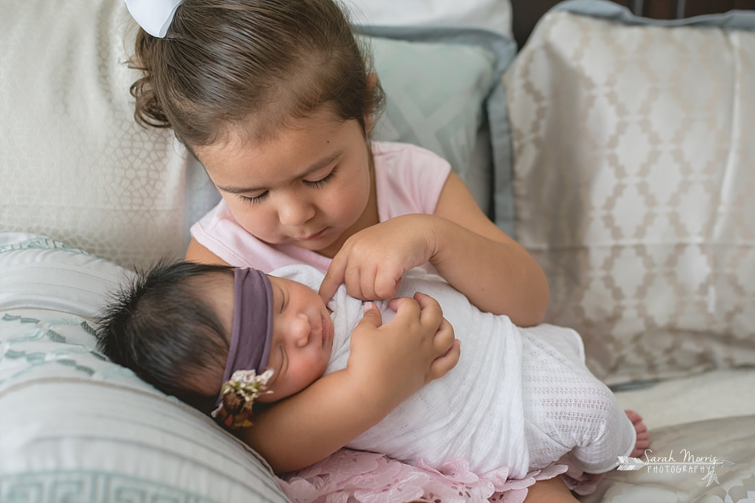 Big sister holding baby sister on bed with newborn baby sister for the lifestyle portion of her newborn photo session