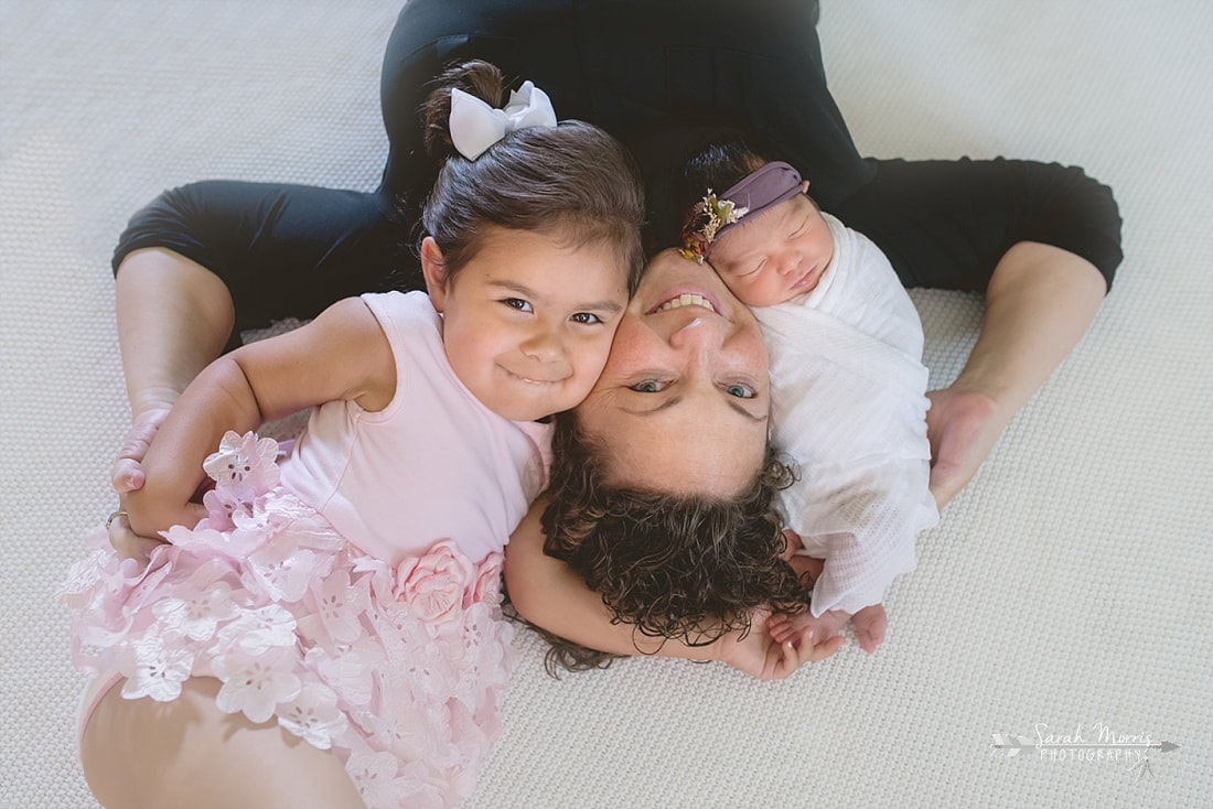 Mother holding both daughters on white blanket during the lifestyle portion of her newborn photo sessionPicture
