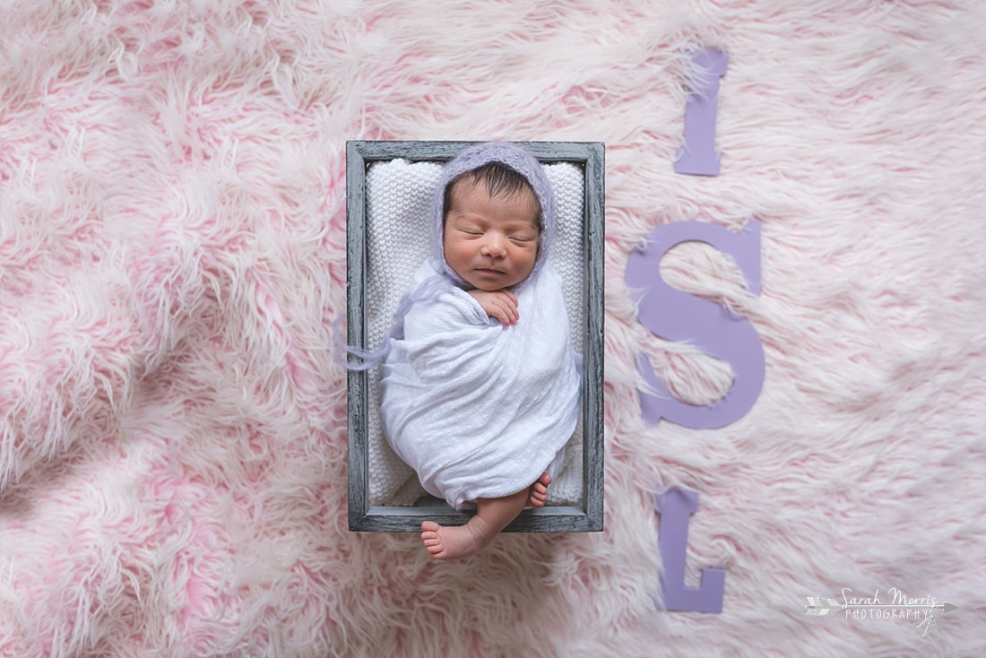 Newborn baby girl swaddled in a white blanket and wearing a beautiful knit bonnet inside of a basket next to her initials on a pink fur blanket during the posed portion of her newborn session