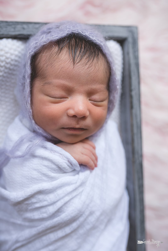 Newborn baby girl swaddled in a white blanket and wearing a beautiful knit bonnet inside of a basket on a pink fur blanket during the posed portion of her newborn session