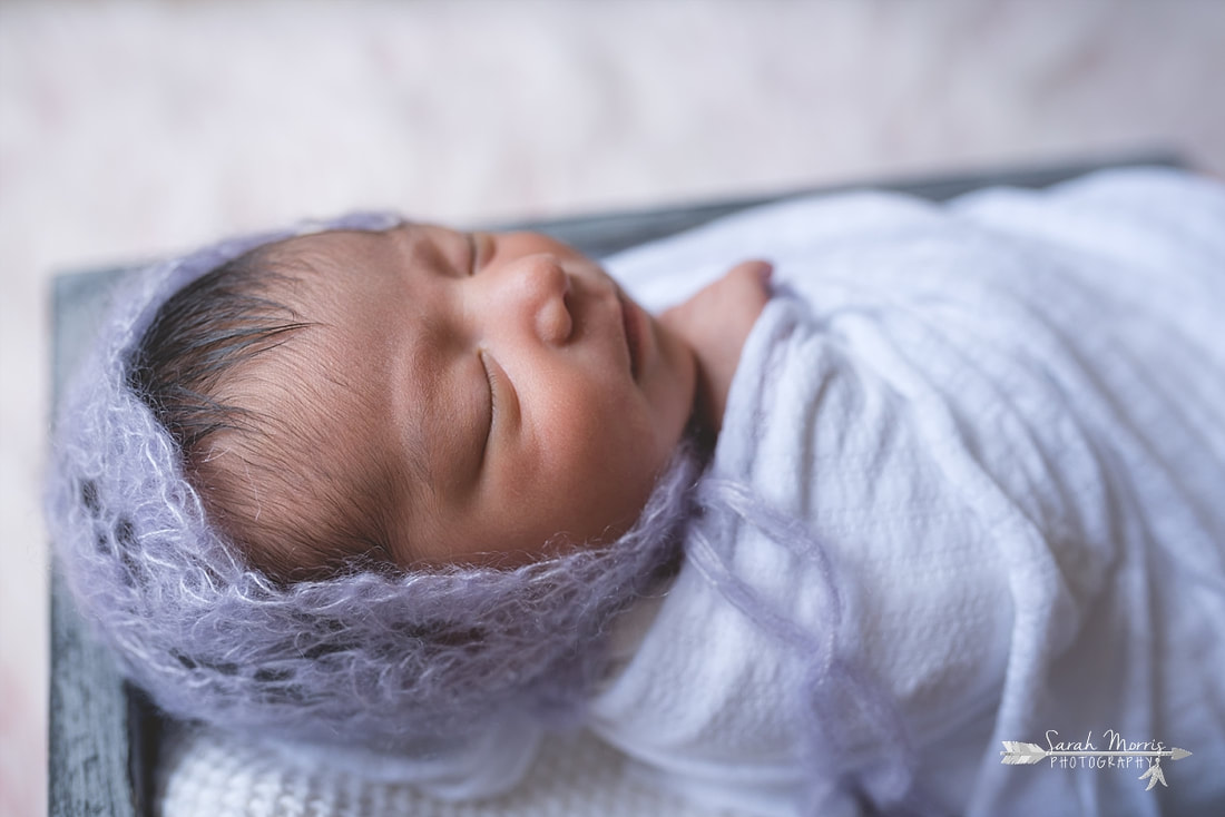 Newborn baby girl swaddled in a white blanket and wearing a beautiful knit bonnet inside of a basket on a pink fur blanket during the posed portion of her newborn session