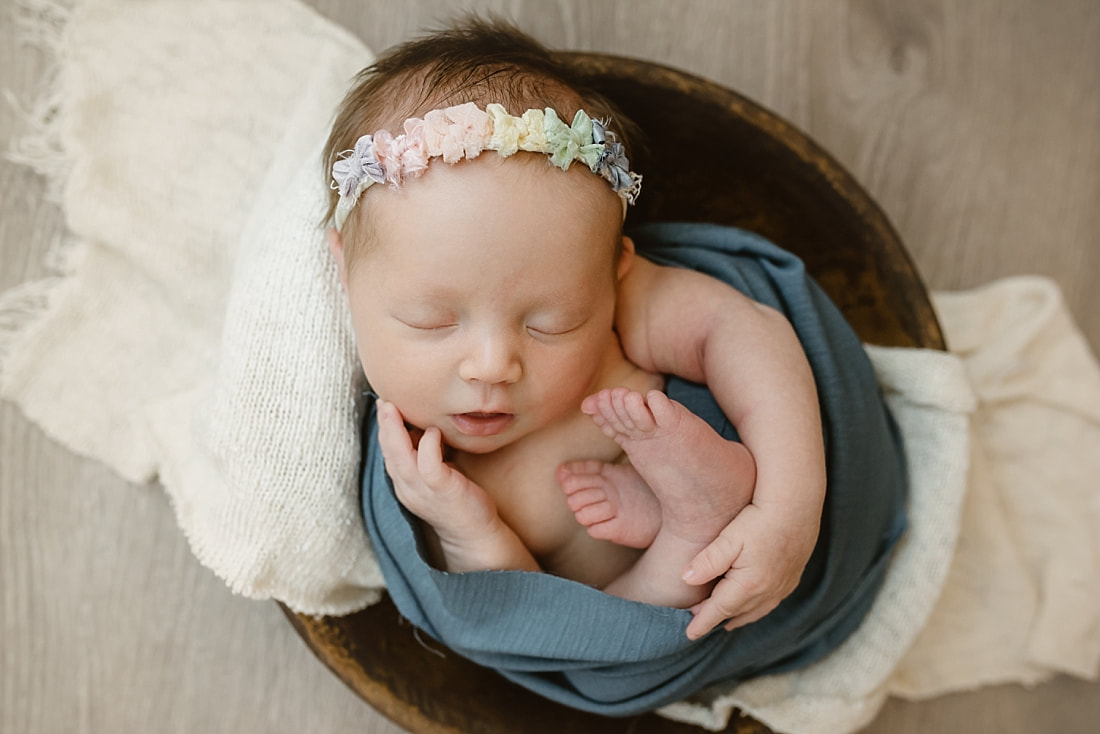 Baby girl wearing a rainbow headband swaddled in blue, sleeping in wooden bowl for newborn photoshoot in Memphis, TN