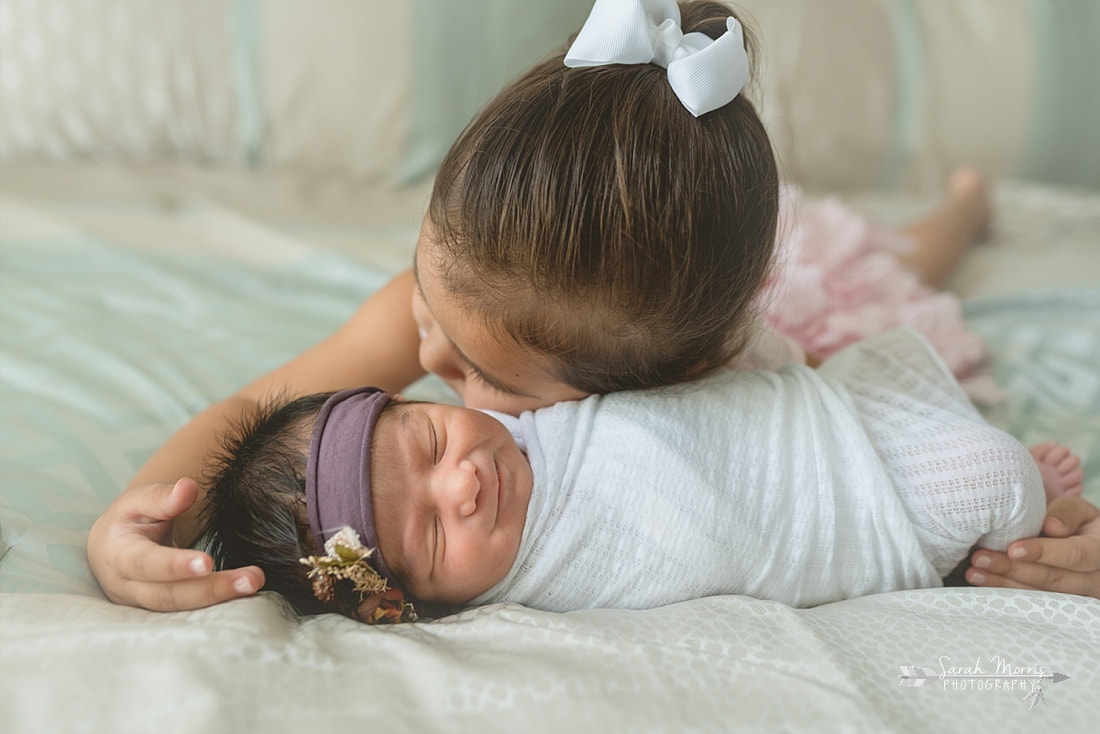 Big sister holding baby sister on bed with newborn baby sister for the lifestyle portion of her newborn photo session