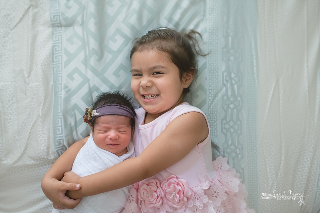 Big sister holding baby sister on bed with newborn baby sister for the lifestyle portion of her newborn photo session