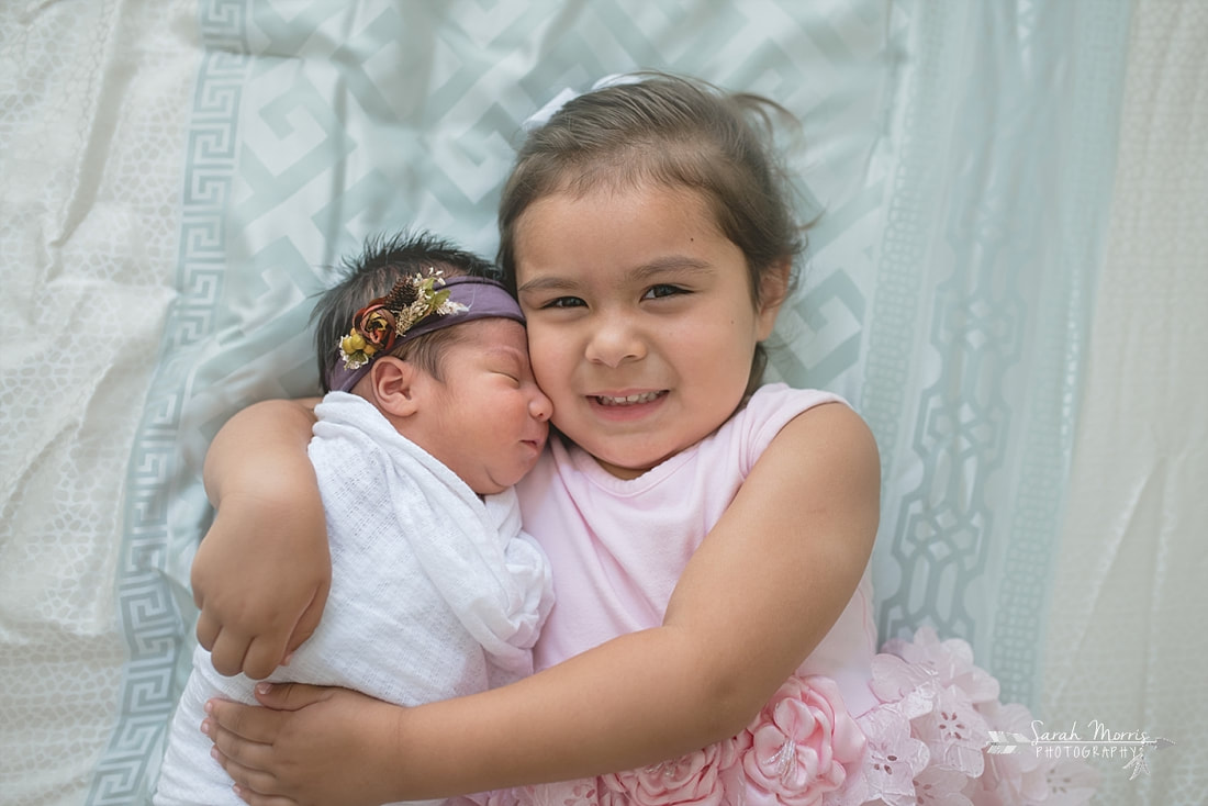 Big sister holding baby sister on bed with newborn baby sister for the lifestyle portion of her newborn photo session