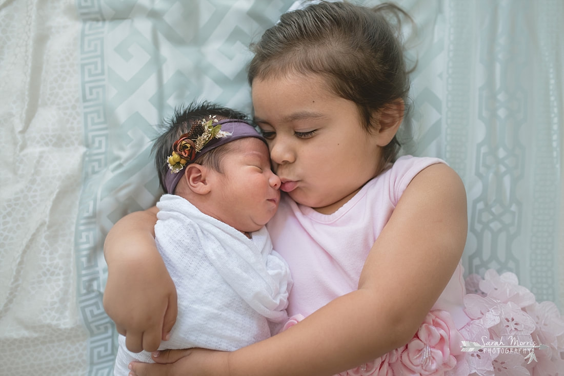 Big sister holding baby sister on bed with newborn baby sister for the lifestyle portion of her newborn photo session