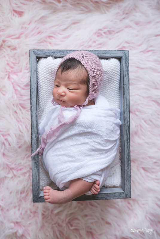 Newborn baby girl swaddled in a white blanket and wearing a beautiful knit bonnet inside of a basket on a pink fur blanket during the posed portion of her newborn session