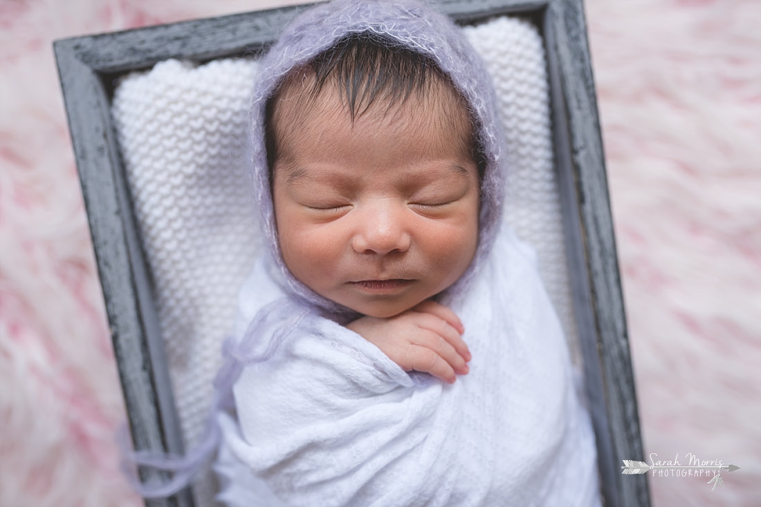 Newborn baby girl swaddled in a white blanket and wearing a beautiful knit bonnet inside of a basket on a pink fur blanket during the posed portion of her newborn session